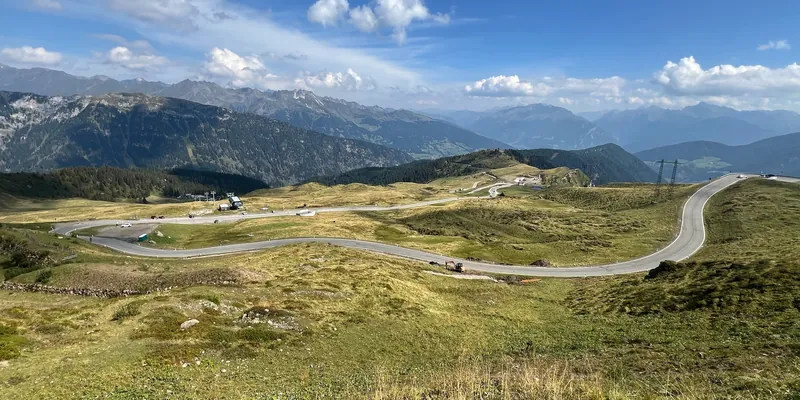 Eine Passstraße schwingt in weiten Bögen durch ein grünes Hochtal, dahinter Bergketten und Schönwetterwolken.