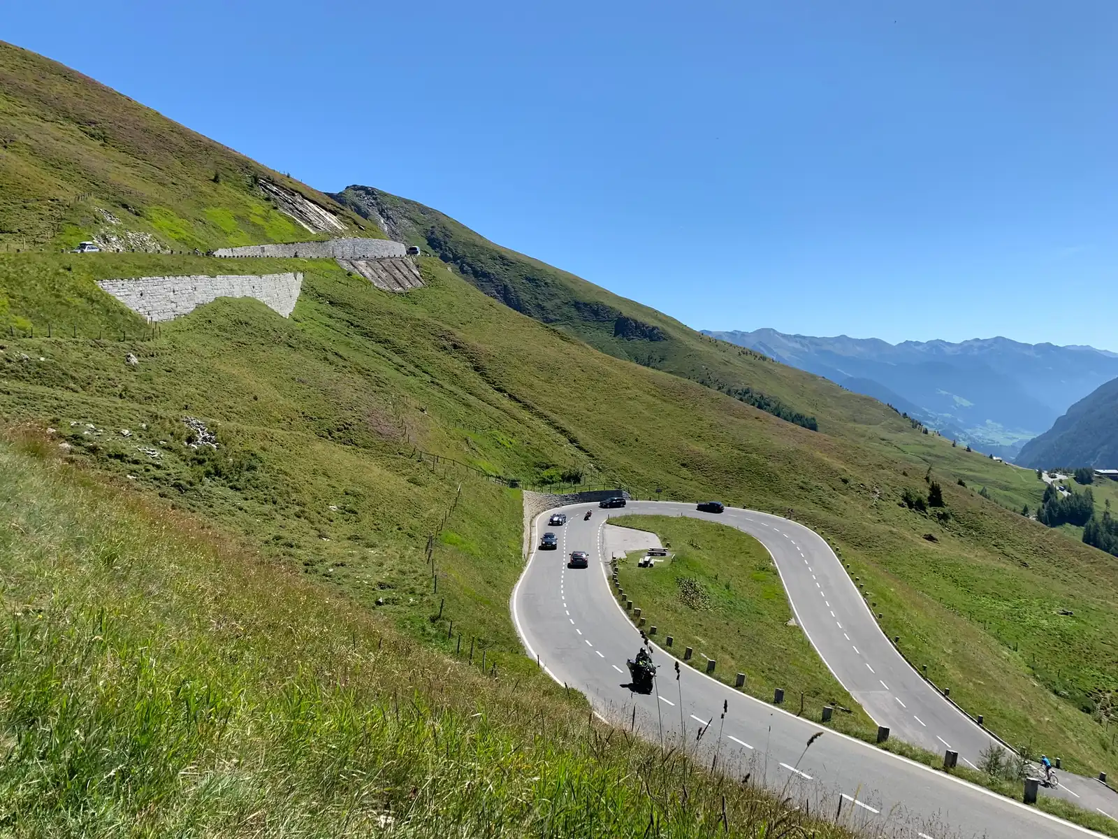 Serpentinen einer Alpenpassstraße ziehen sich durch grüne Bergwiesen, darauf ein paar Motorräder und Autos, dahinter ein weites Tal unter blauem Himmel.