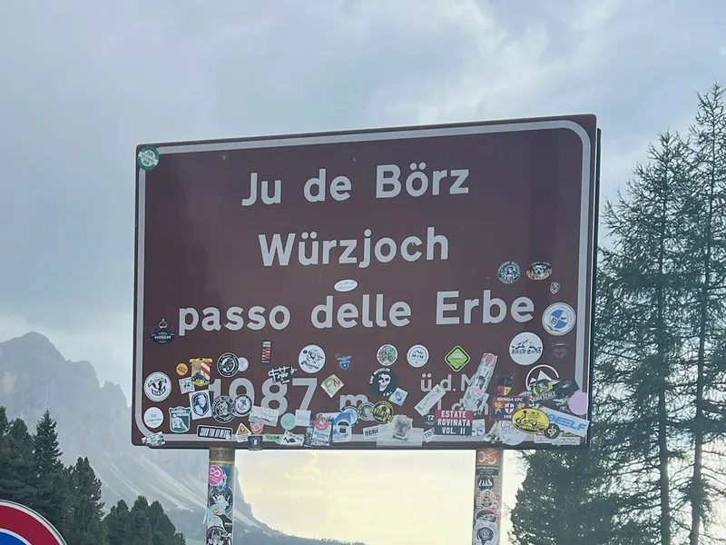 Passschild „Ju de Börz, Würzjoch, Passo delle Erbe, 1987 m“, übersät mit Aufklebern von Motorradfahrern, vor Bergen und Lärchen.